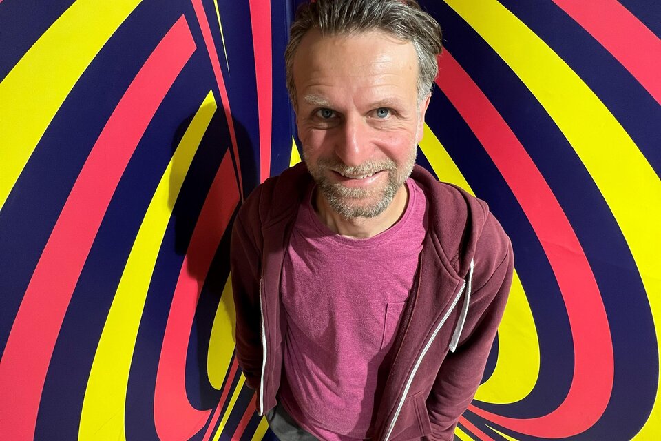 Ein Mann steht vor einem bunten, psychedelischen Hintergrund mit Streifen in Gelb, Blau und Rot. Er lächelt und trägt ein lässiges Outfit. | © © Marva Held