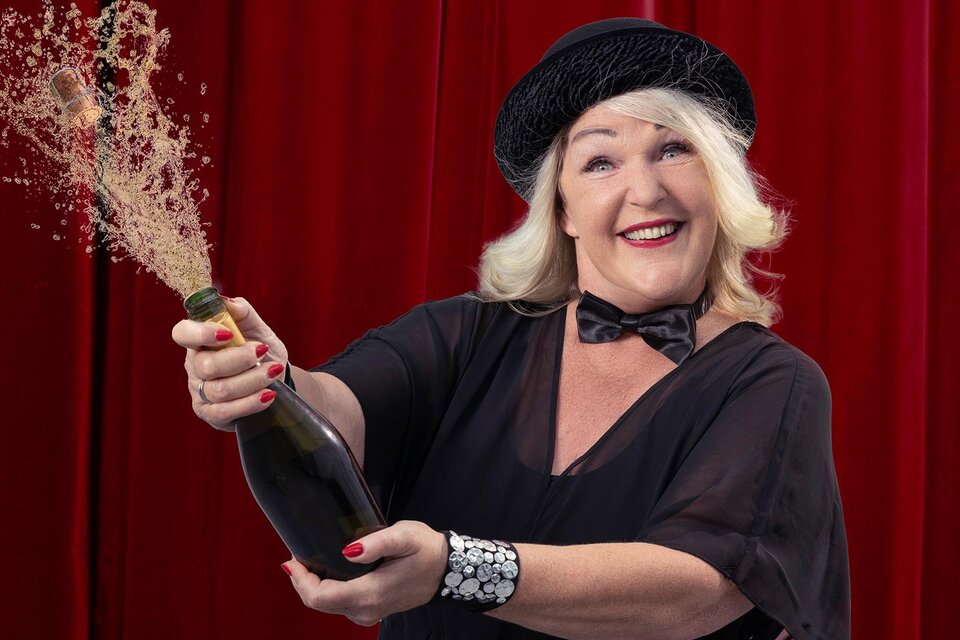 Eine fröhliche Frau in einem eleganten Outfit öffnet eine Flasche Champagner. Im Hintergrund ist ein roter Vorhang zu sehen. | © Barbara Balldini
