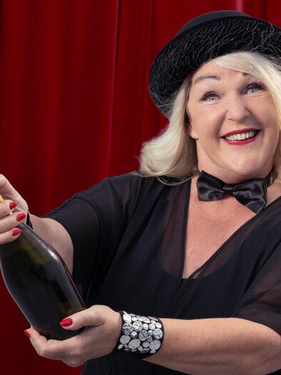 A cheerful woman in an elegant outfit is opening a bottle of champagne. In the background, a red curtain is visible. | © Barbara Balldini