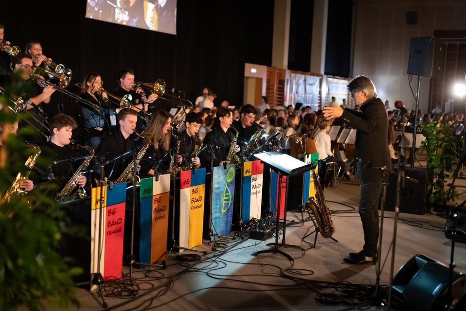 Ein Orchester spielt auf einer Bühne, während der Dirigent dirigiert. Vor dem Orchester stehen bunte Fahnen, die verschiedene Länder repräsentieren. | © Sebastian Friedl