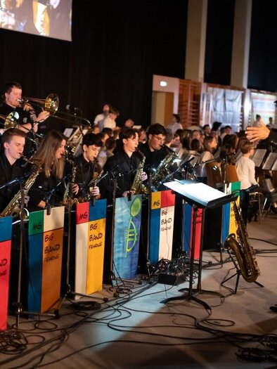 Ein Orchester spielt auf einer Bühne, während der Dirigent dirigiert. Vor dem Orchester stehen bunte Fahnen, die verschiedene Länder repräsentieren. | © Sebastian Friedl