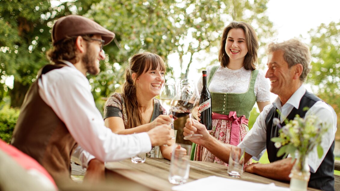Eine fröhliche Gruppe von vier Personen, die an einem Tisch sitzt und auf einander anstößt. Alle sind traditionell gekleidet und genießen die gesellige Atmosphäre im Freien.