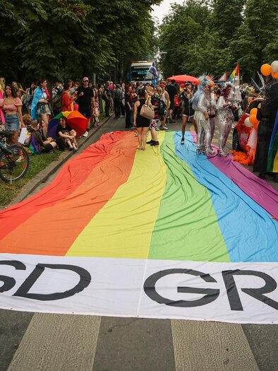 A colorful parade for Christopher Street Day with a large rainbow banner. Many people are celebrating and wearing colorful clothing. | © @Seabstian Neugebauer