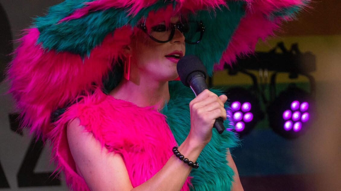 Eine Person trägt ein auffälliges, pinkes und grünes Kostüm mit einem großen Hut und Sonnenbrille. Sie steht am Mikrofon und spricht auf einer Bühne. | © @Sebastian Neugebauer