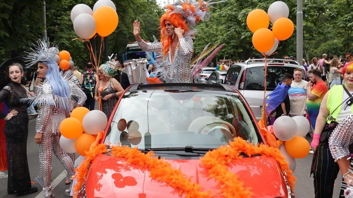 Ein festlich dekoriertes Auto mit orangefarbenen Luftballons und bunten Kostümen. Personen tanzen und feiern auf der Straße. | © @Sebastian Neugebauer