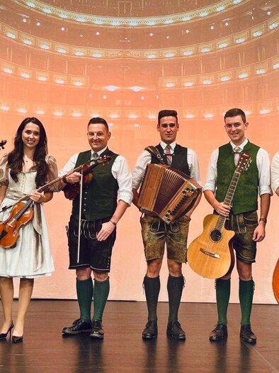 A group of five musicians stands in a room with musical notes on the wall. They are wearing traditional clothing and holding musical instruments in their hands. | © Ausseer Bradlmusi_Bad Aussee_ c Ben Borowskil