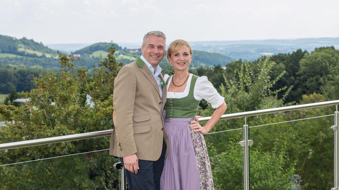Ein Paar in traditioneller Kleidung steht auf einem Balkon mit Blick auf eine grüne Landschaft. Die Atmosphäre wirkt festlich und freundlich. | © Hotel RETTER