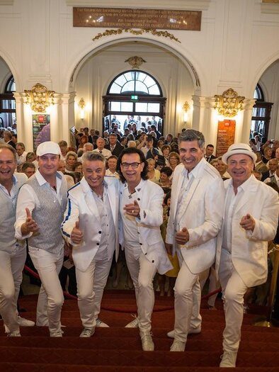 Eine Gruppe von Männern in weißen Anzügen steht auf einer Treppe und lächelt in die Kamera. Im Hintergrund ist eine Menge von Menschen in einem festlichen Raum zu sehen.