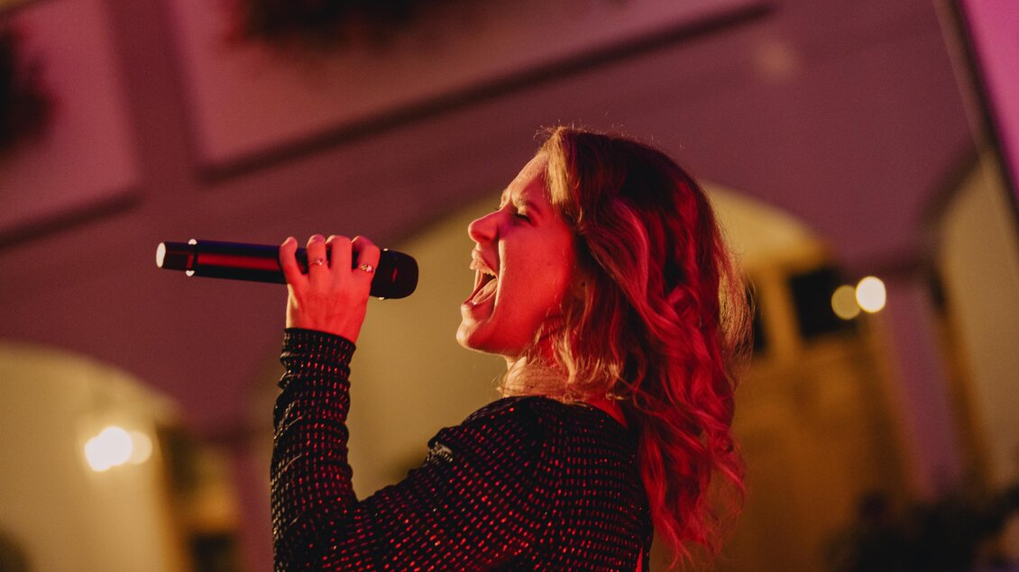 Eine Sängerin performt mit einem Mikrofon in der Hand. Die Bühnenbeleuchtung schafft eine lebendige Atmosphäre.