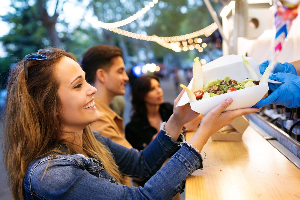 Eine lächelnde Frau erhält eine Portion Essen von einem Food-Truck. Im Hintergrund sind andere Personen zu sehen, die ebenfalls die Atmosphäre genießen. | © European Street Food Festival