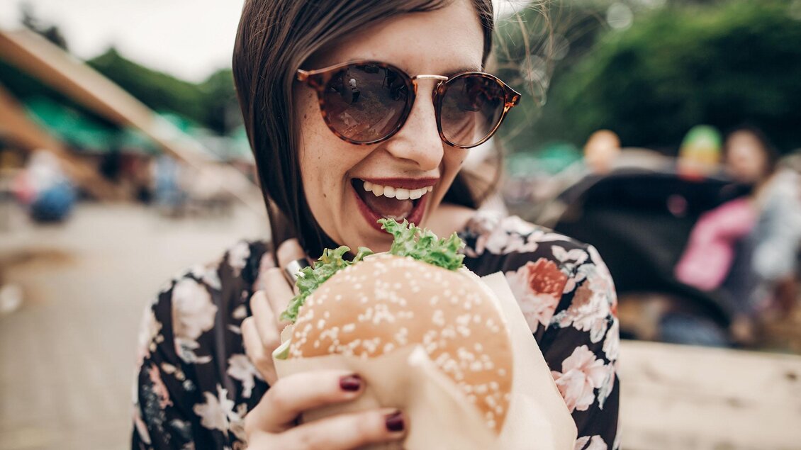 Eine lächelnde Frau mit Sonnenbrille hält einen großen Burger in der Hand. Im Hintergrund sind verschwommen Menschen und eine Freiluftumgebung zu sehen. | © EuropeanStreet Food