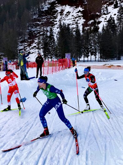 Three cross-country skiers in a competition on a snowy track. In the background, trees and a building can be seen. | © NAZ Eisenerz