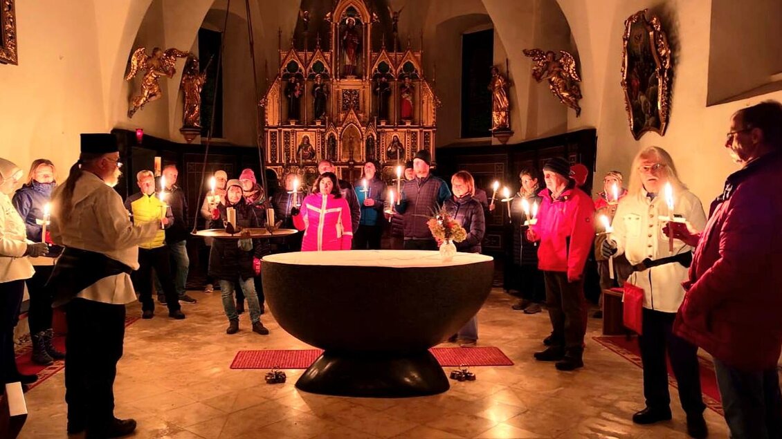 Eine Gruppe von Menschen steht in einer Kirche bei Kerzenlicht. Sie versammeln sich um ein großes Taufbecken im Mittelpunkt des Raumes. | © Hannes Pfandl