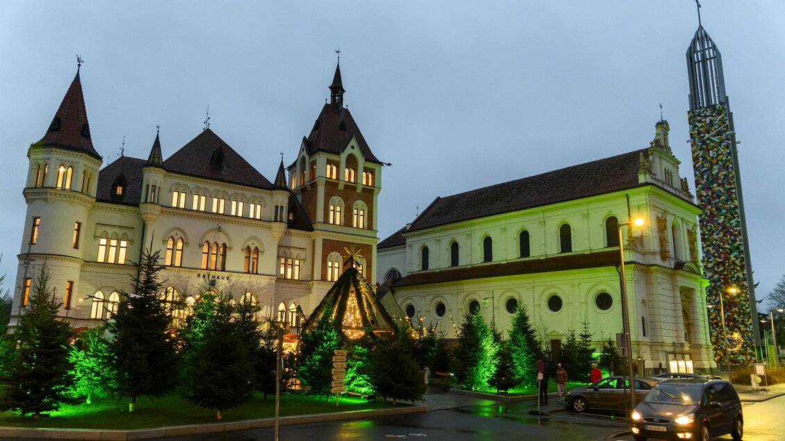 Ein historisches Gebäude mit Türmen und einem modernen Kirchturm. Der Bereich ist festlich mit grünen Lichtern und Tannenbäumen geschmückt. | © Stadtgemeinde Feldbach