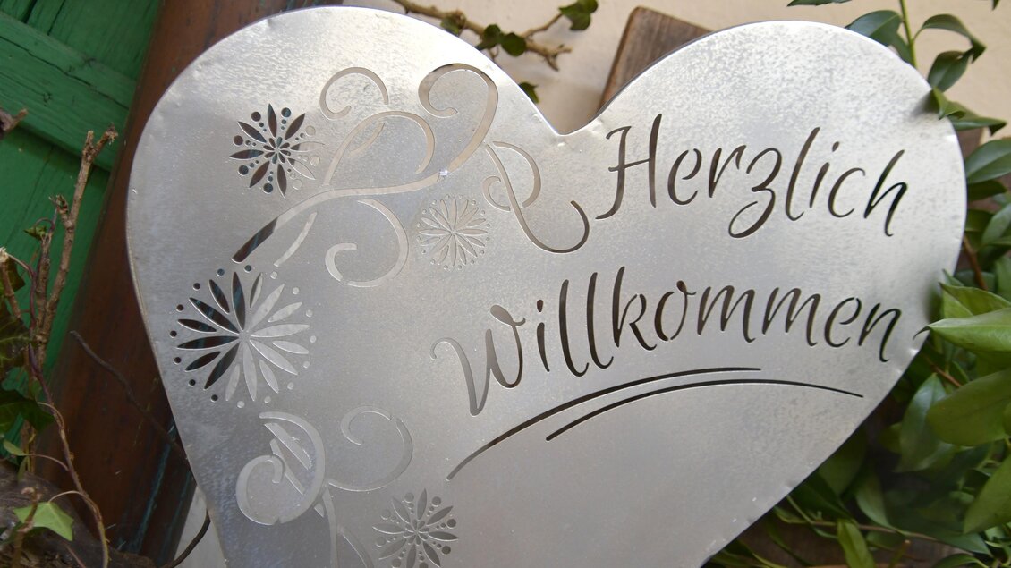 Ein herzförmiges Metallschild mit der Aufschrift "Herzlich Willkommen". Es ist verziert mit filigranen Mustern und Pflanzen. | © Thermen- & Vulkanland