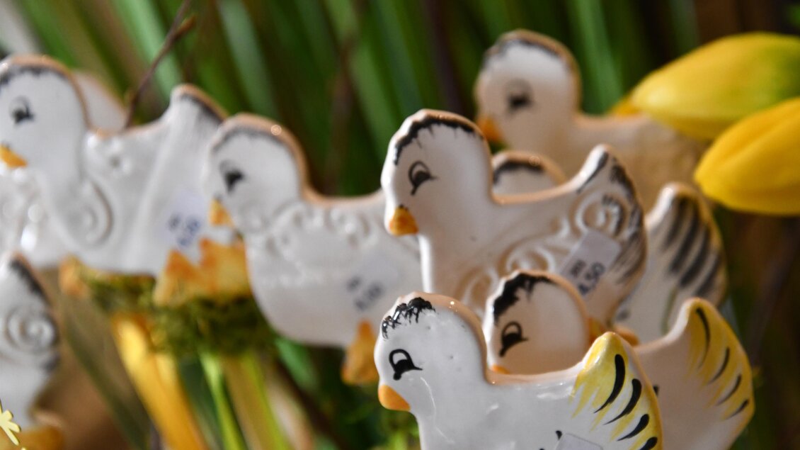 Eine Gruppe von dekorativen, weißen Keramikenten mit bunten Akzenten. Im Hintergrund sind grüne Pflanzen und gelbe Blumen sichtbar. | © Thermen- & Vulkanland