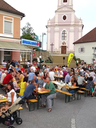 A bustling street scene with many people sitting at long tables and eating. In the background, a church can be seen. | © Gästekonzert_Oststeiermark