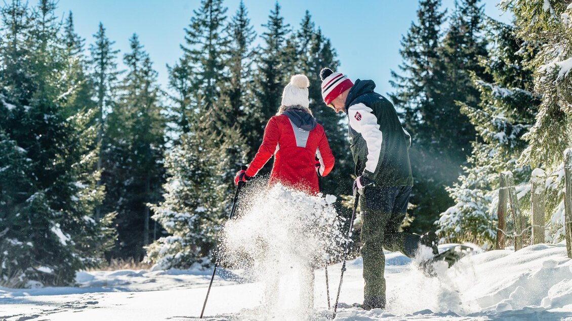 Ein Paar wandert im Schnee, umgeben von hohen Tannenbäumen. Die Sonne scheint und es ist eine klare Winterlandschaft. | © Region Graz-Mias Photoart