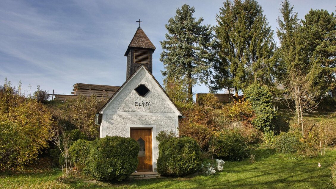 Kleine weiß verputzte Kapelle mit Holztür und Glockenturm am Waldrand. | © WEGES