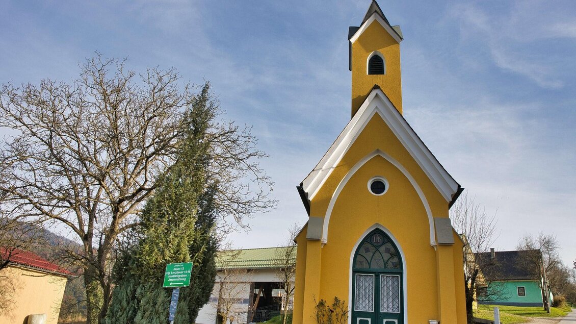Gelb verputzte Kapelle mit Turm und grünem Tor entlang des Wanderwegs. | © WEGES