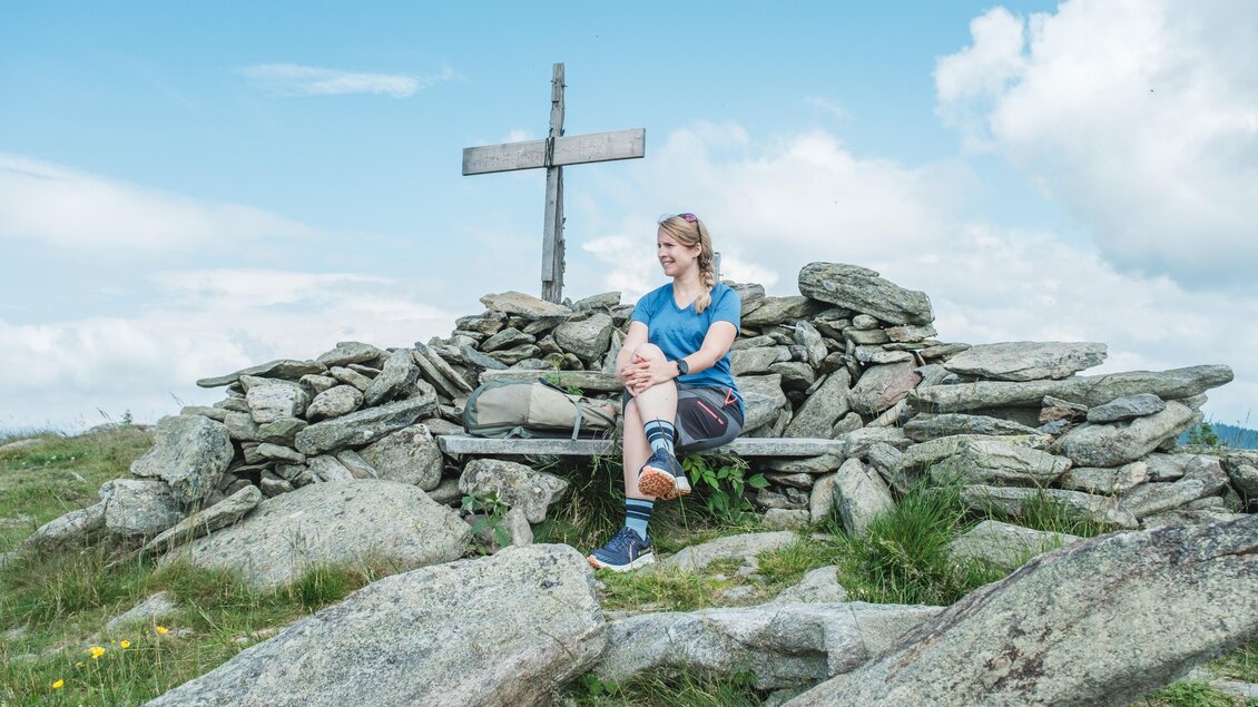 Frau sitzt am Gipfelkreuz der Wieseralm vor Steinmauer mit Rucksack. | © Janine Wenzel
