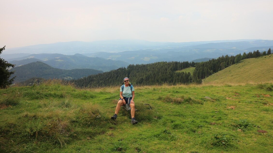 Wanderin sitzt am Walzkogel mit Blick über bewaldete Hügelketten der Weststeiermark bis zum Horizont. | © Heinz Kaltschmidt