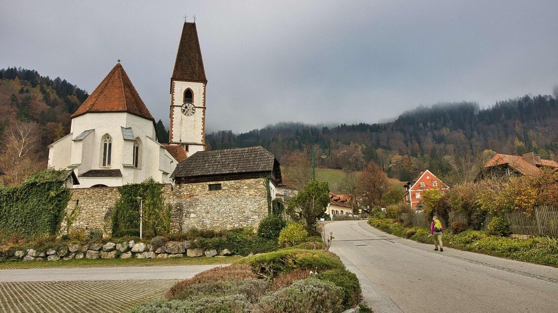 Wanderin geht durch Übelbach, im Vordergrund die Pfarrkirche mit rotem Turmdach. | © WEGES