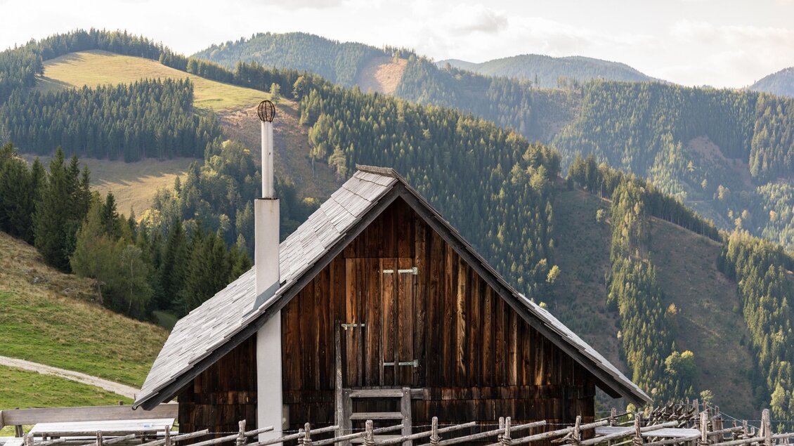 Holzverkleidete Almhütte mit Kamin auf der Gmoaalm vor bewaldeter Hügellandschaft. | © Region Graz-Mias Photoart