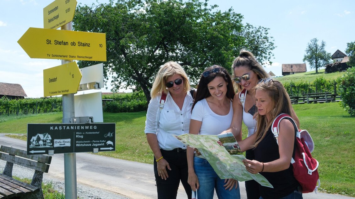 Eine Gruppe von vier Frauen, die eine Karte in einem malerischen Landschaftsbereich betrachten. Im Hintergrund sind Wegweiser und Bäume zu sehen. | © CDM