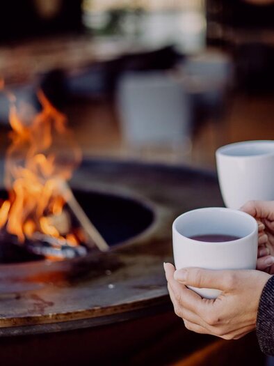 Zwei Personen halten Tassen mit heißem Getränk über einem offenen Feuer. Im Hintergrund sind verschwommene Stühle und ein gemütlicher Raum zu sehen. | © Ratscher Landhaus