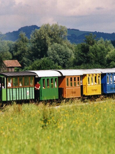 An old steam locomotive with colorful carriages travels through a green meadow. In the background, gentle hills and a cloudy sky can be seen. | © TV Südsteiermark - Riffel Helmuth
