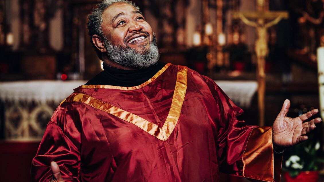 Ein fröhlicher Mann in einer roten Talar steht in einer Kirche. Er hat einen Bart und schaut lächelnd nach oben. | © Karin Gruber