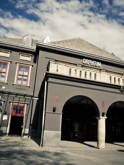 Ein historisches Gebäude mit der Aufschrift "Orpheum" an der Fassade. Die Struktur hat einen eleganten architektonischen Stil und wird von einem blauen Himmel umrahmt. | © (c) Spielstätten - Lupi Spuma