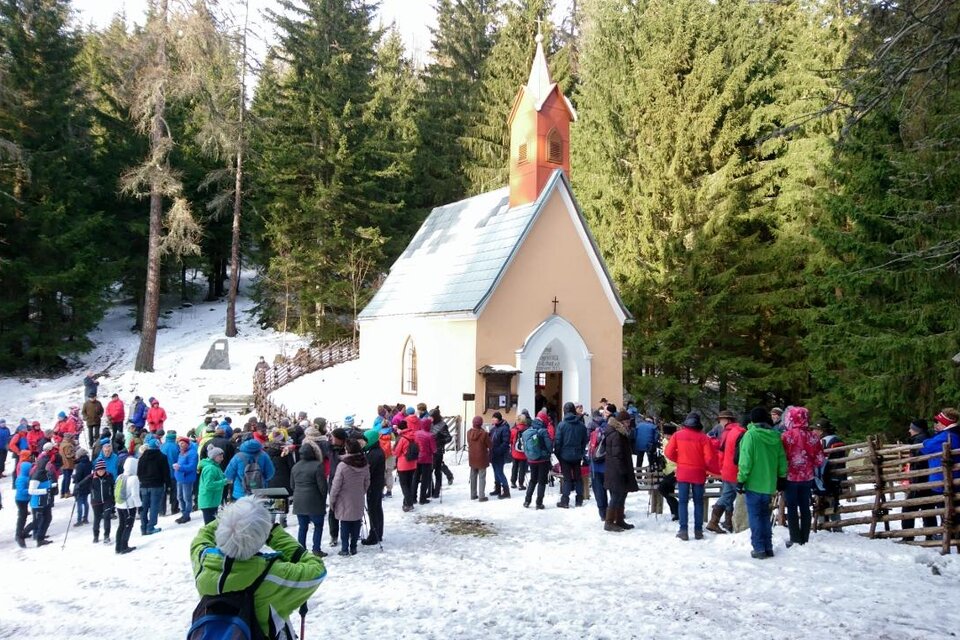 Eine Gruppe von Menschen versammelt sich vor einer kleinen Kirche im Schnee. Im Hintergrund sind hohe, grüne Bäume sichtbar. | © Karin Schäffer