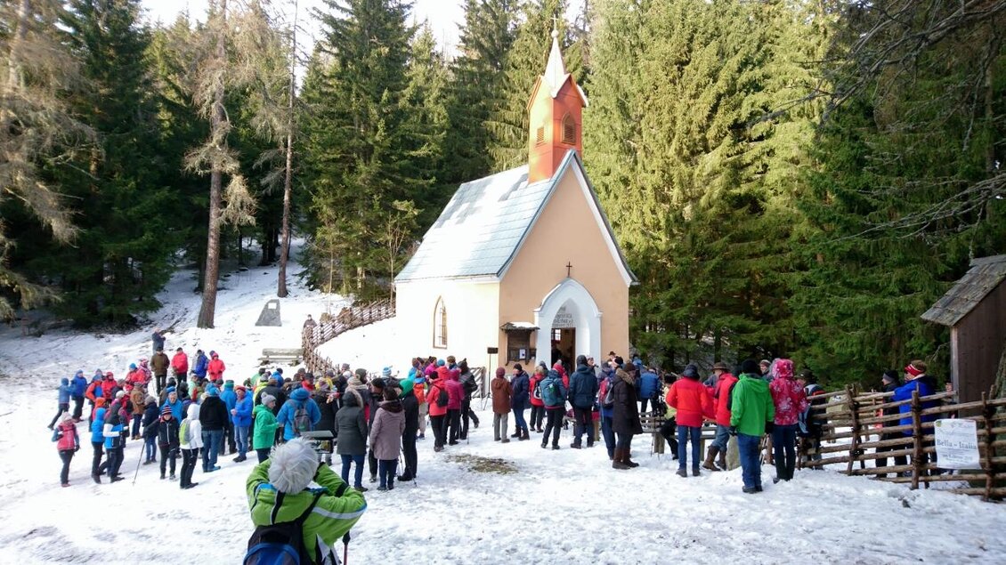 Eine Gruppe von Menschen versammelt sich vor einer kleinen Kirche im Schnee. Im Hintergrund sind hohe, grüne Bäume sichtbar. | © Karin Schäffer