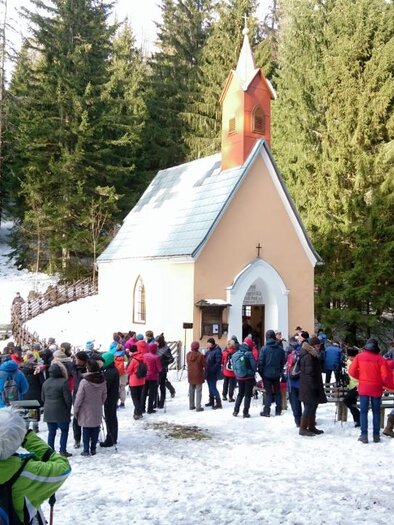 A group of people gathers in front of a small church in the snow. In the background, tall green trees are visible. | © Karin Schäffer