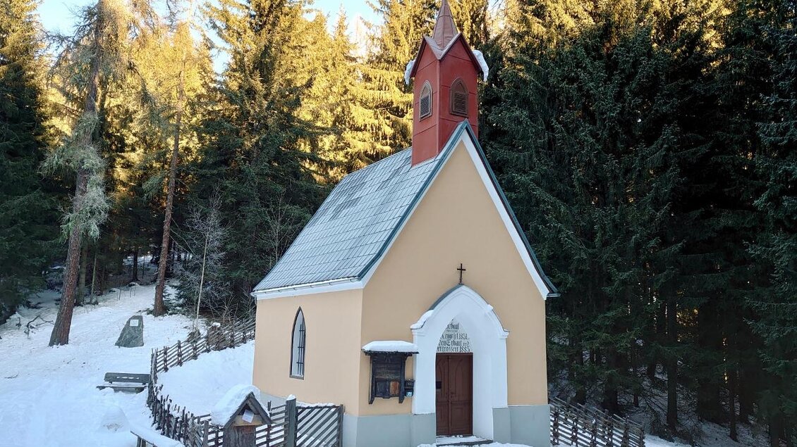 Eine kleine Kirche steht in einer verschneiten Landschaft, umgeben von dichten Bäumen. Der Himmel ist klar und die Sonne scheint. | © Karin Schäffer