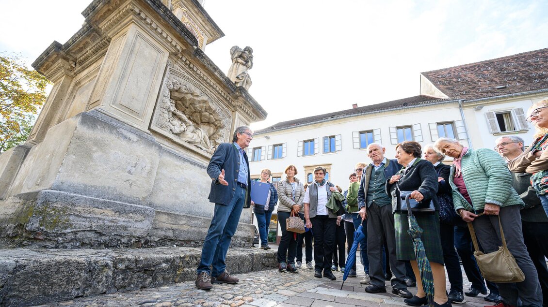 Eine Gruppe von Menschen hört einem Stadtführer vor einem historischen Denkmal zu. Es ist ein sonniger Tag und die Teilnehmer sind neugierig und aufmerksam. | © TV Thermen- & Vulkanland