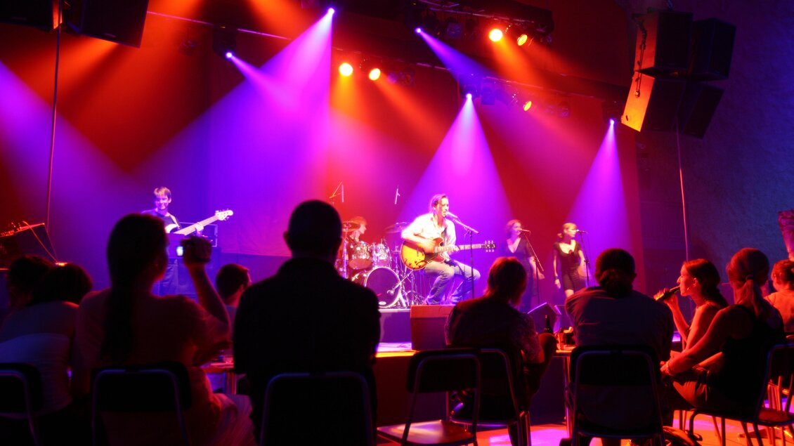 Eine Live-Band spielt auf der Bühne mit bunten Lichtern. Das Publikum sitzt in Stühlen und genießt die Musik. | © (c)grazer_spielstätten