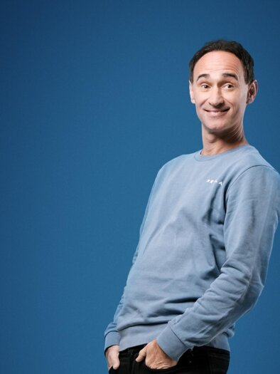 Porträt von Gernot Kulis, einem österreichischen Komiker, der in einem blauen Sweatshirt vor einem blauen Hintergrund steht. In einer entspannten Pose lächelt er charmant in die Kamera. | © Hans Leitner