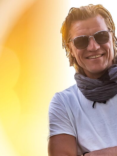 Gery Seidl posiert lächelnd mit Sonnenbrille und Schal vor einem goldenen Hintergrund. | © Johann Eder