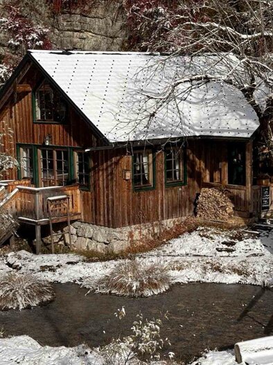 A charming, wooden-clad house in a snow-covered landscape. Surrounding it are bare trees and a small pond visible. | © Cafe Viola, Marina Schobegger