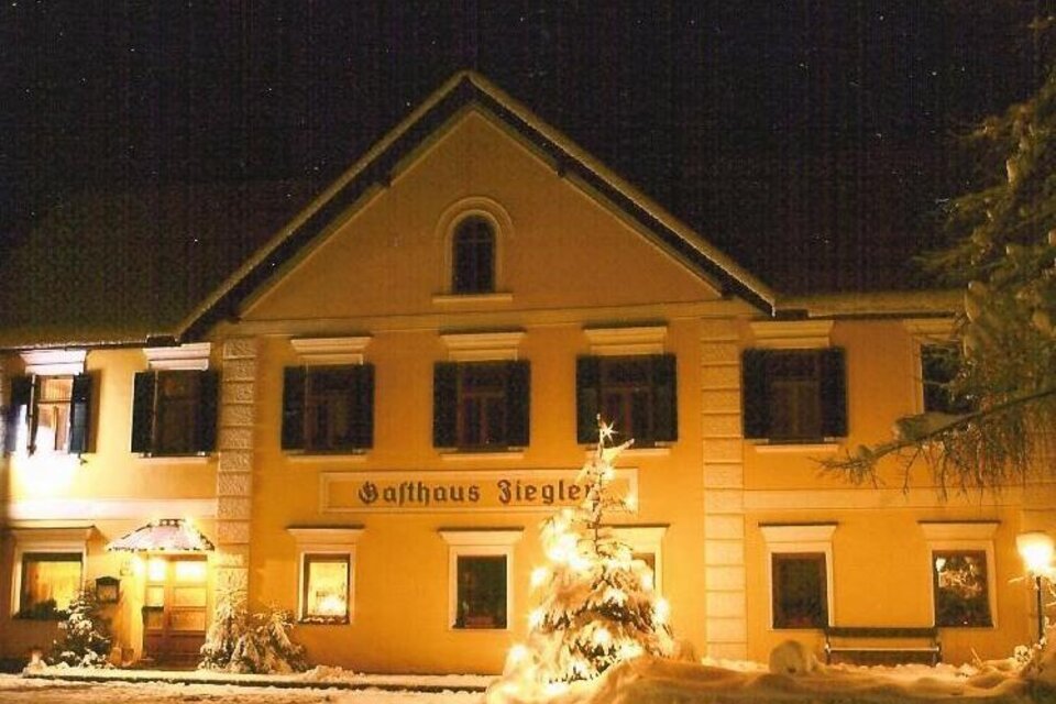 Ein Gemütliches Gasthaus im Winter, beleuchtet von warmem Licht. Schnee bedeckt den Boden und ein festlich geschmückter Weihnachtsbaum steht vor dem Gebäude. | © Seiler-Ziegler