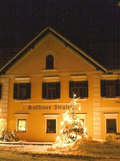 Ein Gemütliches Gasthaus im Winter, beleuchtet von warmem Licht. Schnee bedeckt den Boden und ein festlich geschmückter Weihnachtsbaum steht vor dem Gebäude. | © Seiler-Ziegler