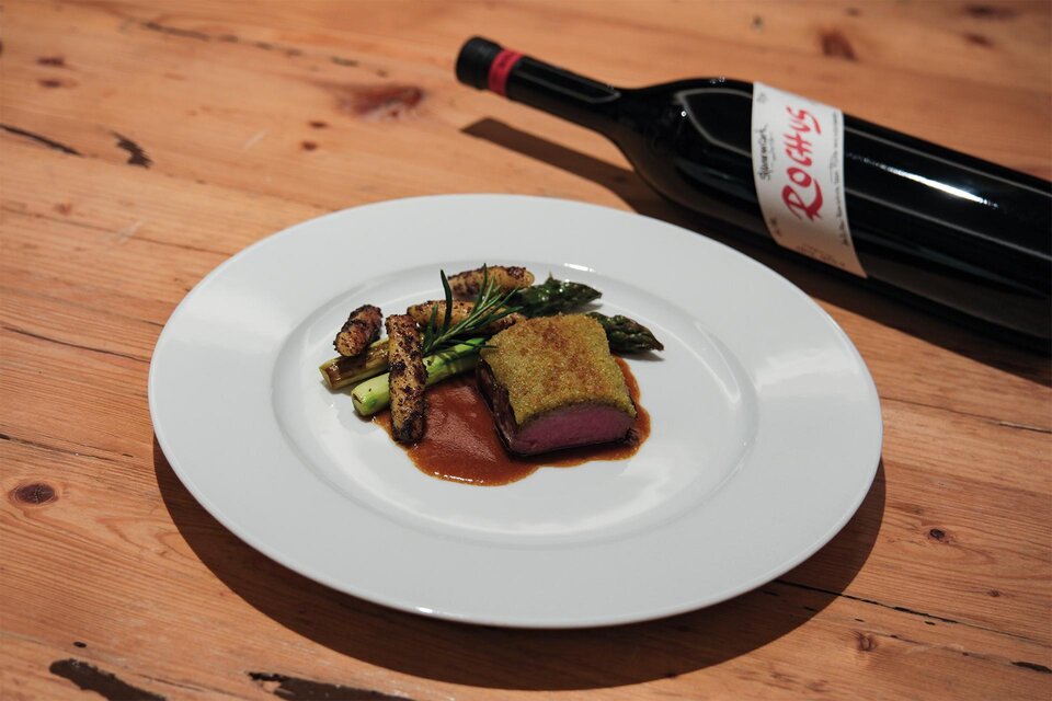 Ein Teller mit gebratenem Fleisch, umgeben von Gemüse und einer Soße. Daneben steht eine Flasche Rotwein auf einem Holztisch. | © Weinschloss Thaller