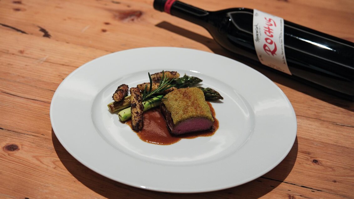 Ein Teller mit gebratenem Fleisch, umgeben von Gemüse und einer Soße. Daneben steht eine Flasche Rotwein auf einem Holztisch. | © Weinschloss Thaller