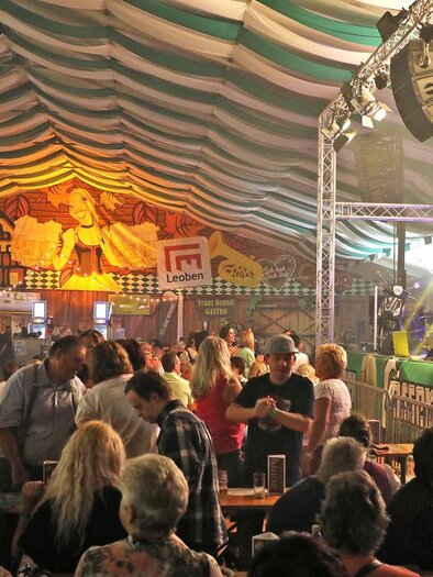 A festive event in a heated tent with people dancing and celebrating. Musicians perform on stage while the atmosphere is lively and cheerful. | © Wiesenfest