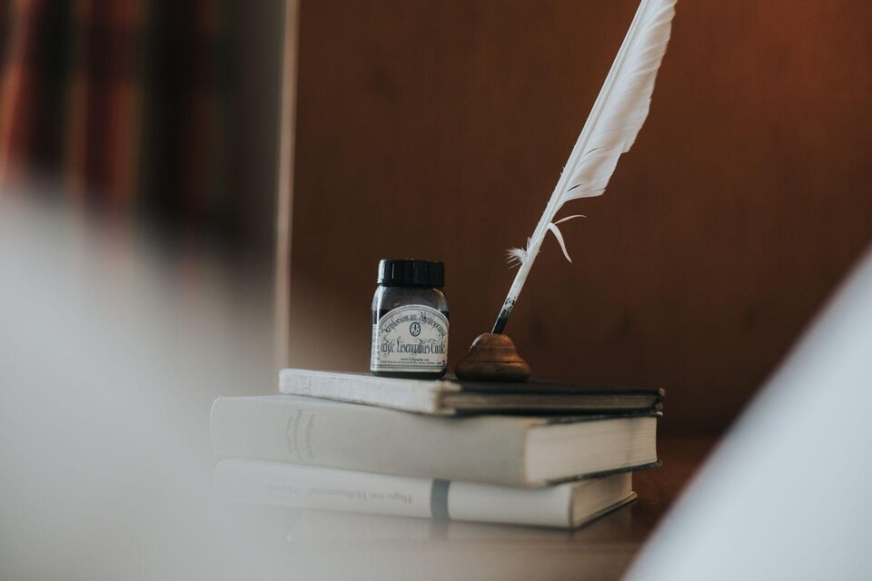 Ein Federkiel und ein Tintenfass stehen auf einem Stapel Bücher. Im Hintergrund sind sanfte Farben und eine warme Atmosphäre zu sehen. | © Buch (c) Wasnerin, Bad Aussee