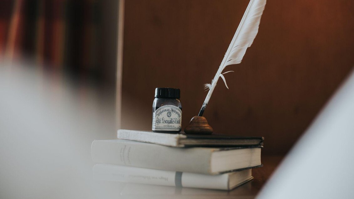 Ein Federkiel und ein Tintenfass stehen auf einem Stapel Bücher. Im Hintergrund sind sanfte Farben und eine warme Atmosphäre zu sehen. | © Buch (c) Wasnerin, Bad Aussee