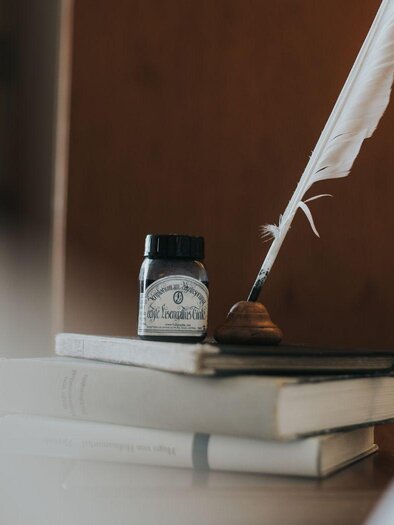 A quill and an inkpot are on a stack of books. In the background, gentle colors and a warm atmosphere can be seen. | © Buch (c) Wasnerin, Bad Aussee